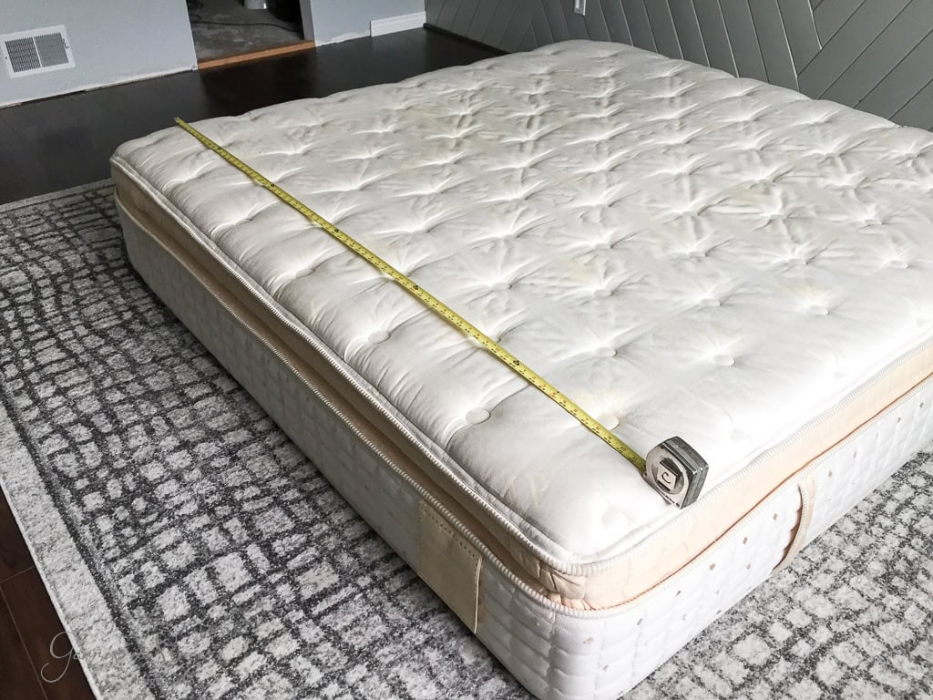A white mattress on a gray patterned rug with a yellow measuring tape stretched across it, showing its width. The setting is a dimly lit room with a wooden floor and a herringbone patterned wall.