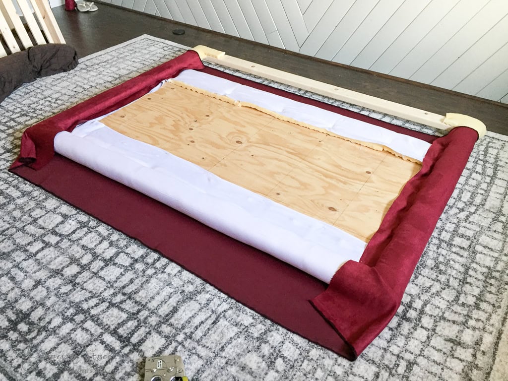 A DIY fabric headboard in progress, laid out on a table. The headboard frame is covered with red fabric, partially folded and secured over a wooden board. A staple gun is visible in the corner.