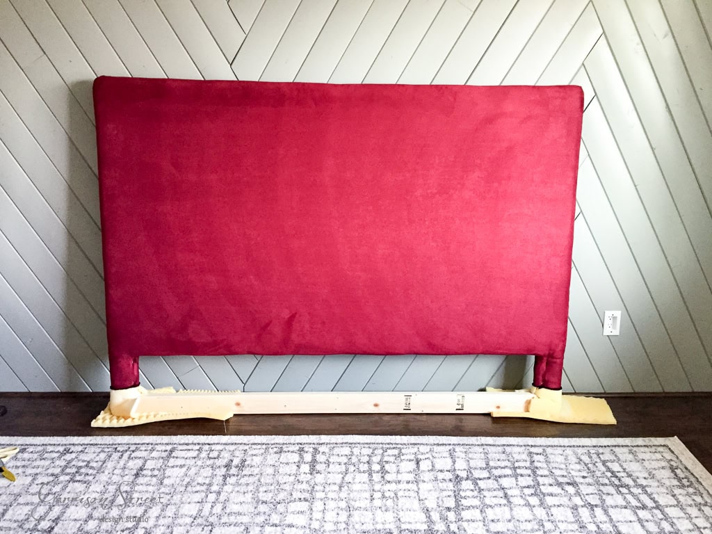 A large red upholstered headboard leans against a chevron-patterned wall. The headboard has foam padding at the bottom corners for protection. A portion of a patterned rug is visible on the floor.