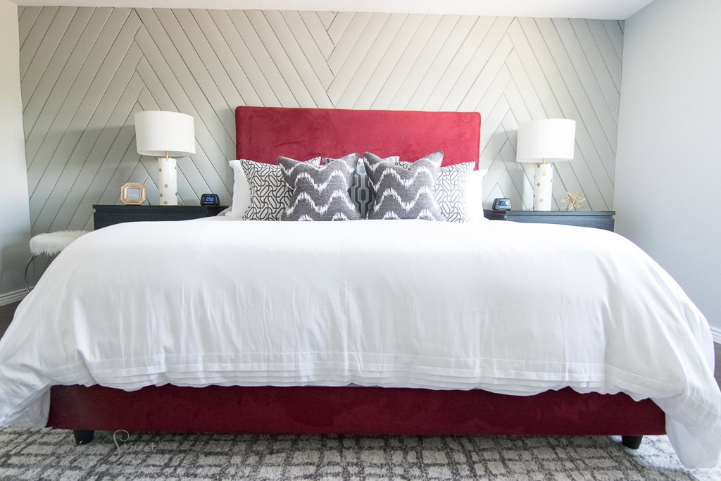 A modern bedroom features a large bed with a red upholstered headboard, white bedding, and decorative pillows. Two matching bedside tables with lamps flank the bed. The wall behind has diagonal wood paneling, and the floor has a patterned rug.