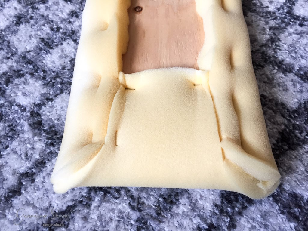 A piece of foam partially wrapped around a rectangular wooden block with visible staples securing the foam. The setup is placed on a textured gray carpet.