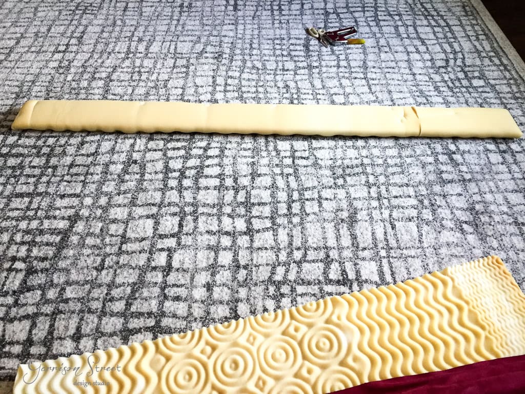 An unrolled, yellow foam topper with a wavy texture is laid out on a patterned rug, stretching from the top to the bottom. A pair of scissors and a pen or marker are placed nearby on the rug.