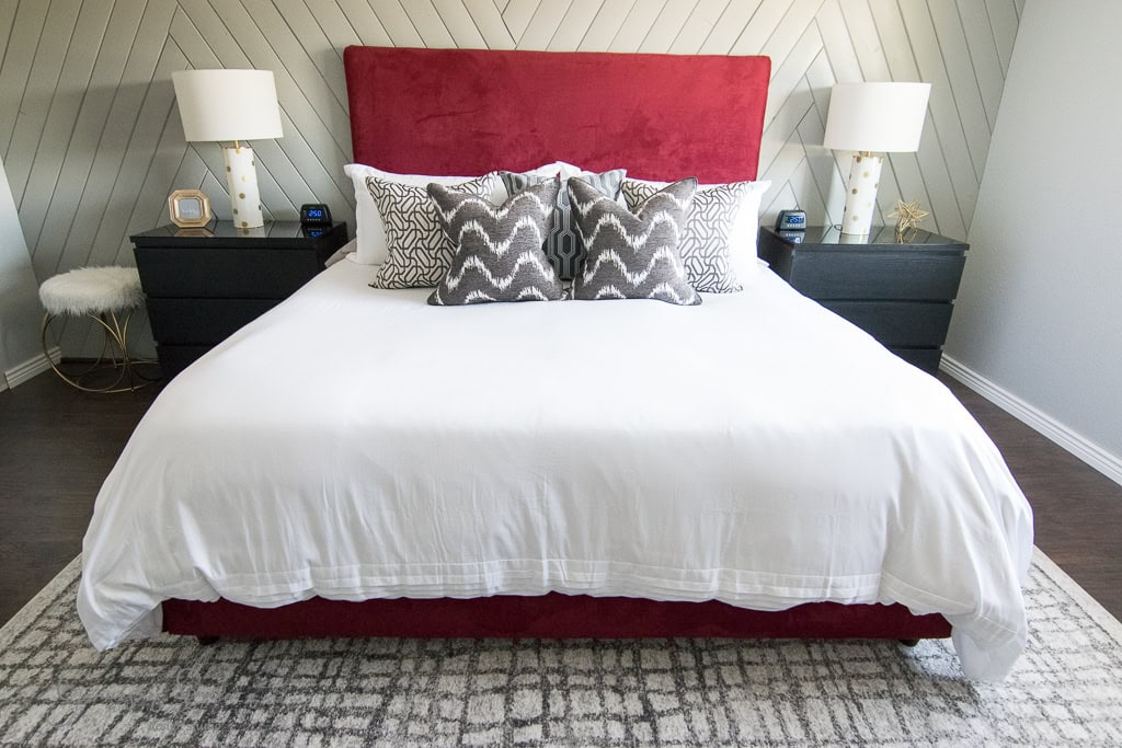 A bedroom with a red headboard and white bedding featuring patterned gray and white pillows. Two black nightstands with white lamps flank the bed. A textured rug covers the floor, and a light wood herringbone wall serves as the backdrop.