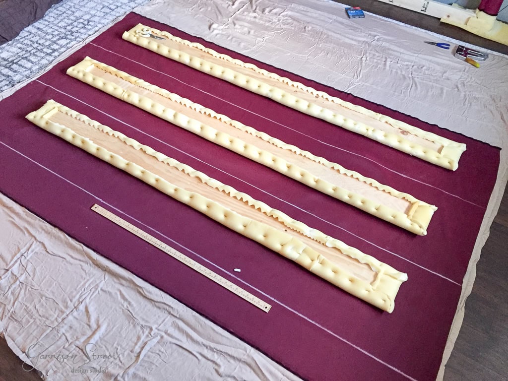Three long, cushioned headboards in a row on a red fabric sheet, with a ruler for scale. Tools and materials are scattered around, indicating an upholstery project in progress.
