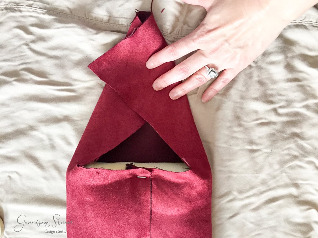 A close-up of a person's hand holding a piece of red leather fabric, folded to form an envelope shape, on a beige surface. The hand has a ring on the ring finger.