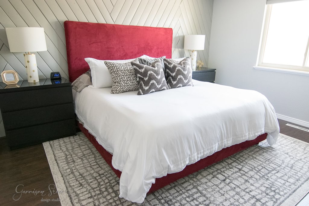 A modern bedroom with a large bed featuring a red headboard and white bedding. The bed is adorned with patterned pillows. Black nightstands flank the bed, each topped with a lamp. A gray patterned rug covers the dark floor.