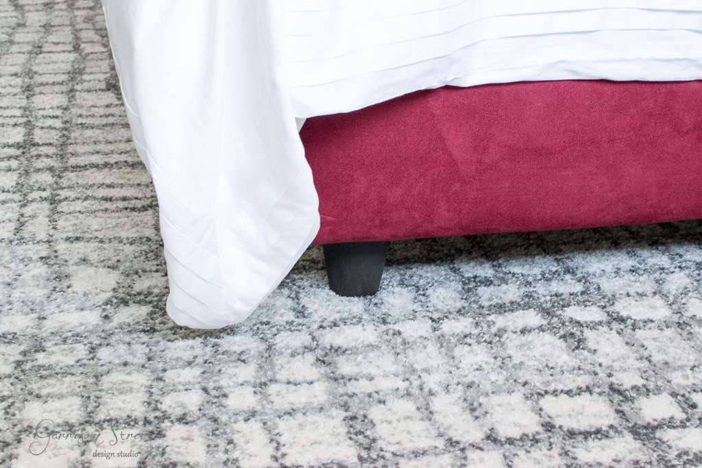 A red bed frame with a white sheet draped over the corner, set on a gray patterned carpet. The bed frame has black legs, and the texture of the carpet features intersecting lines and a grid-like design.