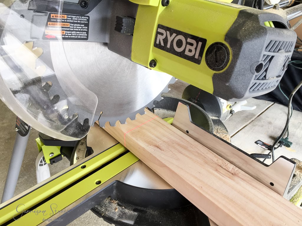 A Ryobi circular saw in use, cutting a wooden plank. The saw is positioned on a work surface, with sawdust scattered around. The saw's metallic blade is visible, and a safety cover is partially lifted.
