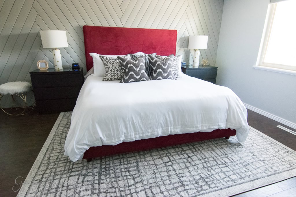 A modern bedroom with a white and gray color scheme features a bed with a red headboard, white bedding, and patterned pillows. Two black nightstands with white lamps flank the bed. A geometric rug lies on the dark wooden floor.