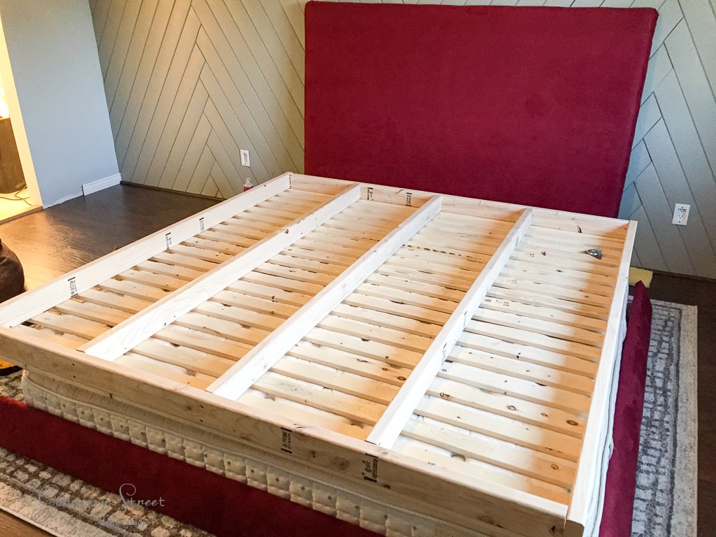 A partly assembled wooden bed frame with vertical slats and a large red upholstered headboard in a room with herringbone-patterned walls and dark wooden floors.