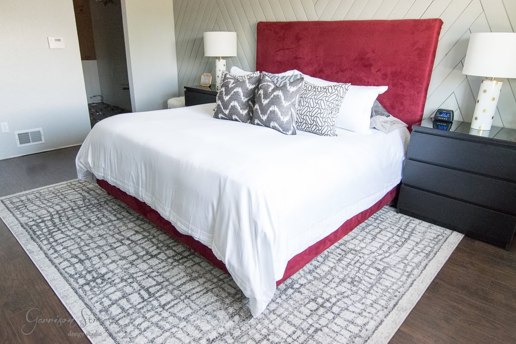 Bedroom with a large bed featuring a red headboard and white bedding. Two gray-patterned pillows rest on the bed. Black nightstands with lamps are on each side. A patterned rug is on the floor, and walls are light gray.
