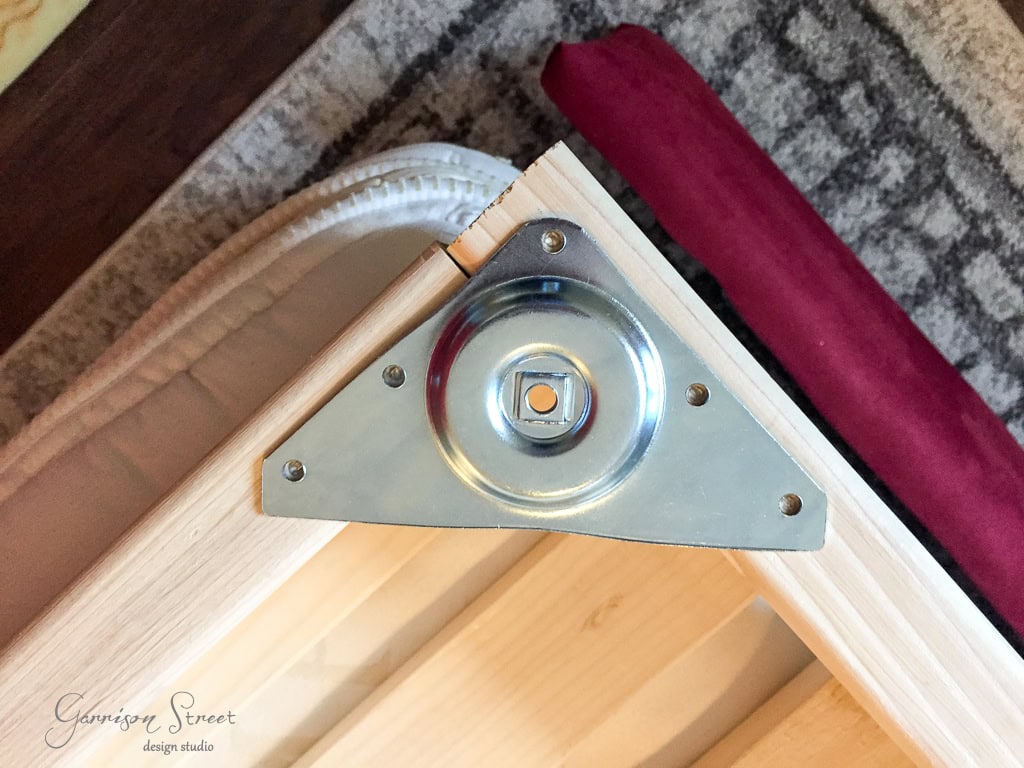 Close-up of a wooden frame corner with a triangular metal bracket featuring a round metal wheel and a bolt. The frame is placed on a textured surface with a glimpse of a dark red mat nearby.