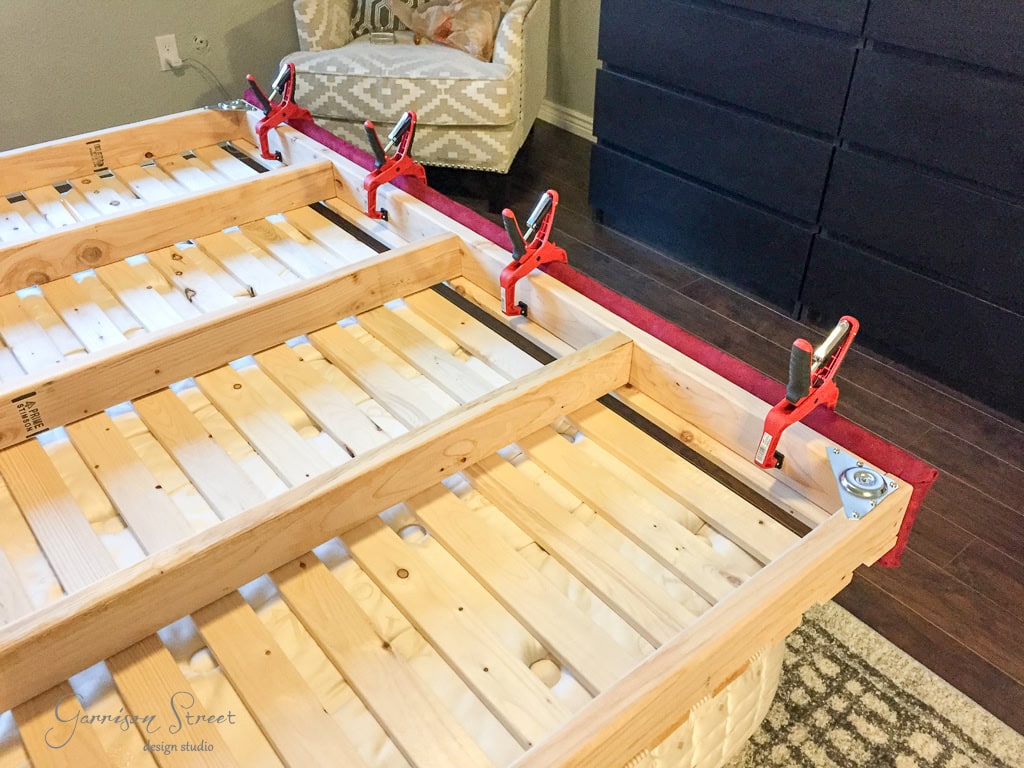 A wooden bed frame under construction, with red clamps securing the rails. The carpentry work is laid out on a patterned rug in a room with an armchair and a dark dresser in the background.