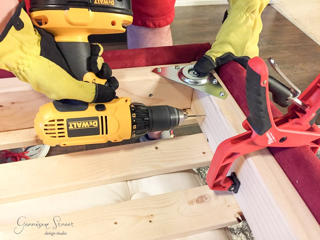 A person wearing yellow gloves uses a yellow power drill on a wooden furniture frame, attaching a metal swivel plate. A red clamp secures the frame. The scene suggests a DIY furniture assembly project.