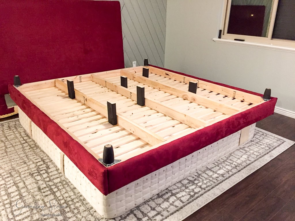 A wooden bed frame with a red upholstered headboard is in a carpeted room. The bed frame, lacking a mattress, has visible wooden slats and black support legs. The room has a window and light-colored walls.