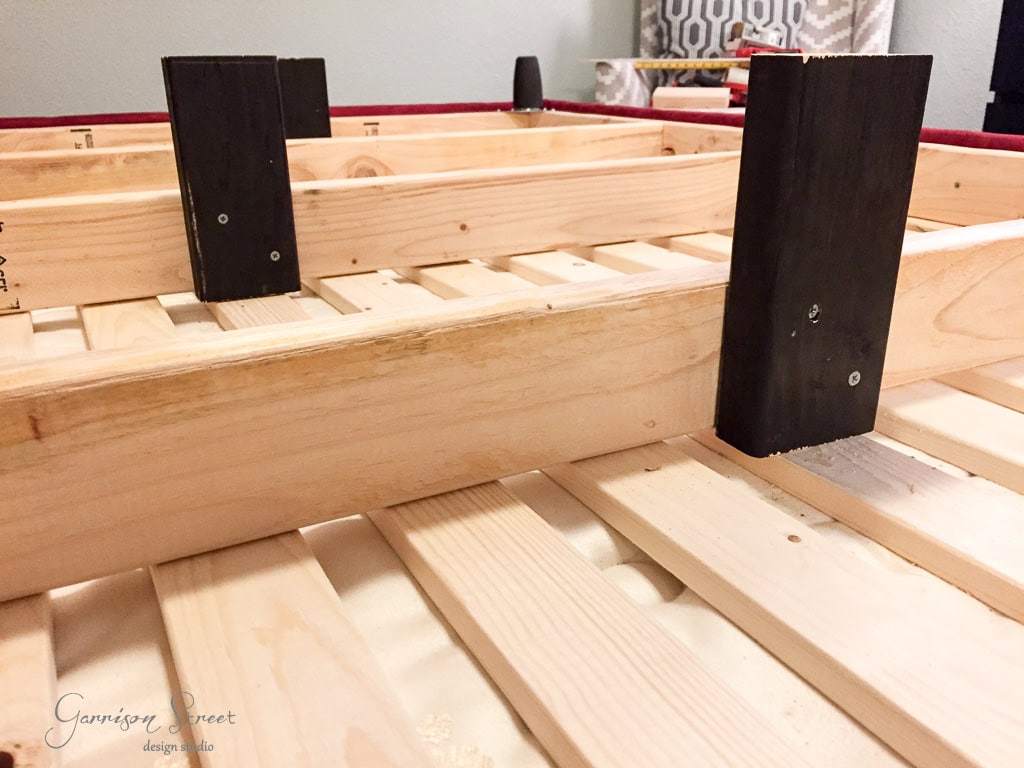 Close-up of a wooden bed frame under construction. The frame features slats and black vertical support beams. Part of a room with a patterned curtain is visible in the background.