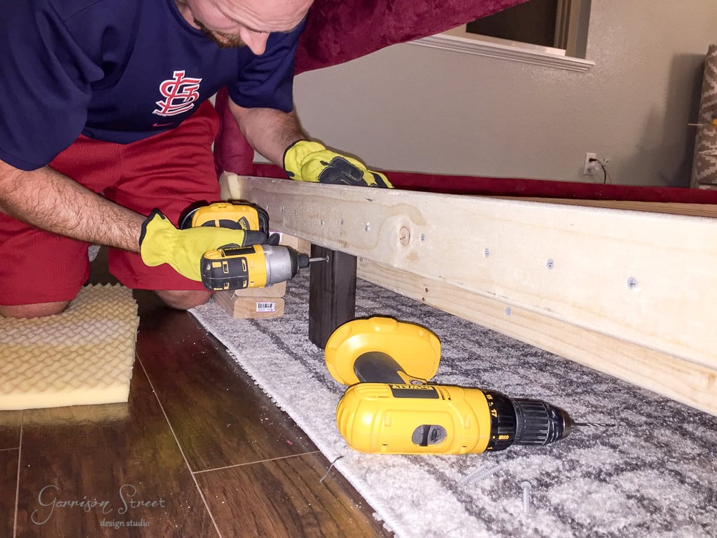 A person in a blue shirt and red shorts is assembling a piece of furniture using a power drill. Wooden planks and tools are visible on a polished wooden floor. They are wearing yellow work gloves.