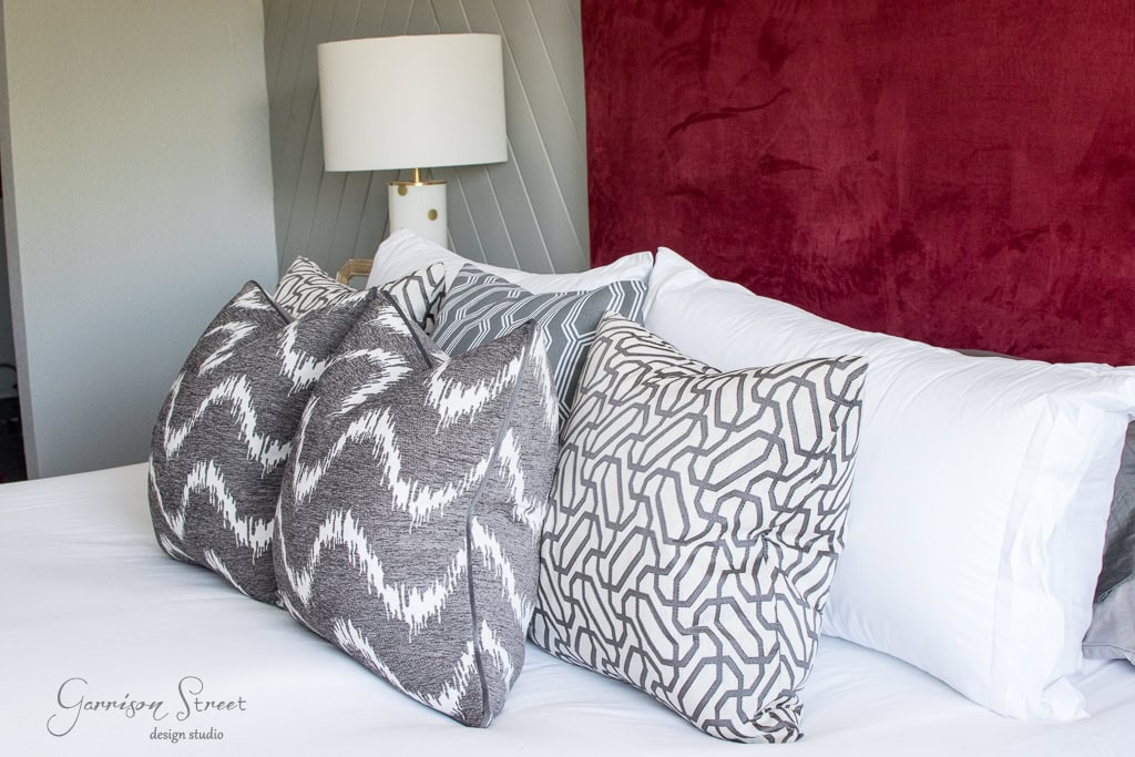 A neatly made bed featuring three patterned throw pillows in gray and white tones. The bed has a white comforter and is set against a textured panel in maroon and gray, with a modern white table lamp beside it.