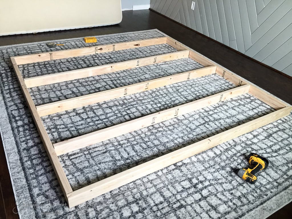 Wooden frame laid on a gray patterned rug, possibly for furniture assembly. A yellow drill and measuring tape are nearby, along with a pencil. Walls feature gray diagonal paneling and a section of dark wooden flooring is visible.