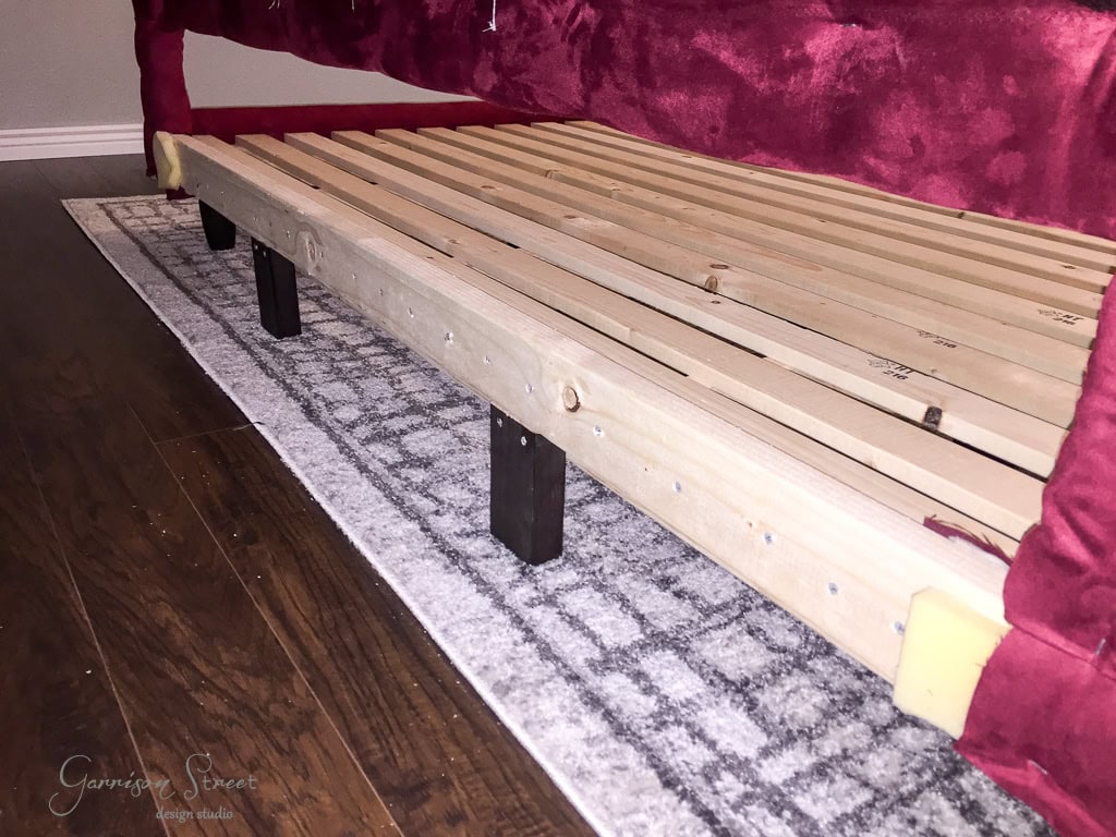 A wooden bed frame with slats, partially covered by a burgundy fabric, sits on a gray patterned rug over hardwood flooring. The frame rests on several black rectangular legs.