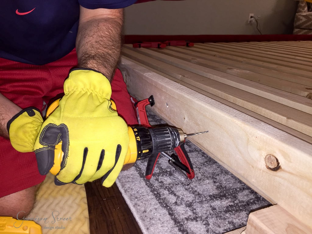 A person wearing yellow gloves uses a cordless drill on a wooden board. The board is clamped to a surface, and the setup appears to be part of a carpentry or DIY project.