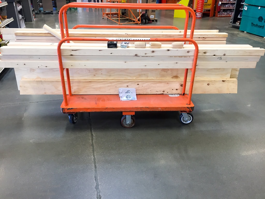 A large orange flatbed cart loaded with various sizes of light-colored wooden planks is positioned on a smooth, gray concrete floor in a hardware store. The background shows aisles with assorted hardware and materials.