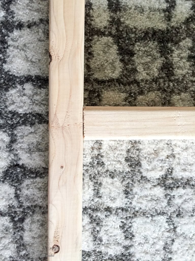 A close-up view of a wooden frame corner positioned on a gray and white patterned carpet. The wood has a natural, light grain texture, and the carpet features a grid-like design.