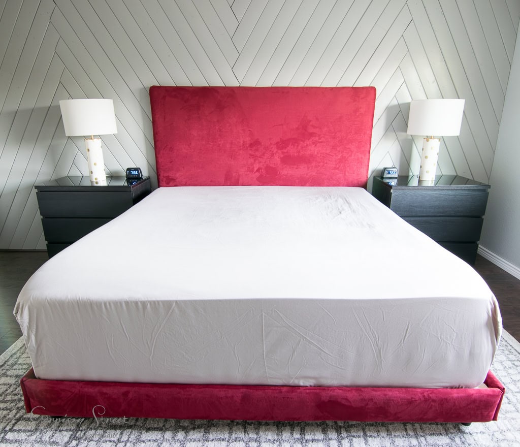A neatly made bed with a red velvet headboard and white bedding is centered between two dark bedside tables, each with a white lamp. The wall features a herringbone pattern, and the floor has a gray patterned rug.