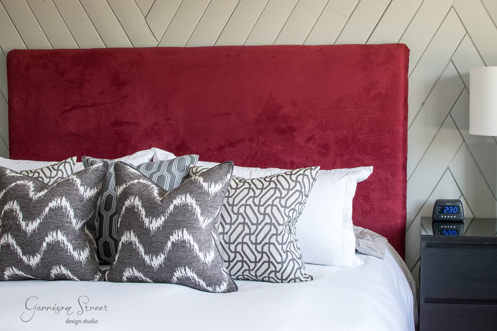A bedroom with a red upholstered headboard, white bed linens, and patterned gray and white throw pillows. A nightstand with a digital alarm clock and lamp is on the right side. The wall features a diagonal wooden pattern.