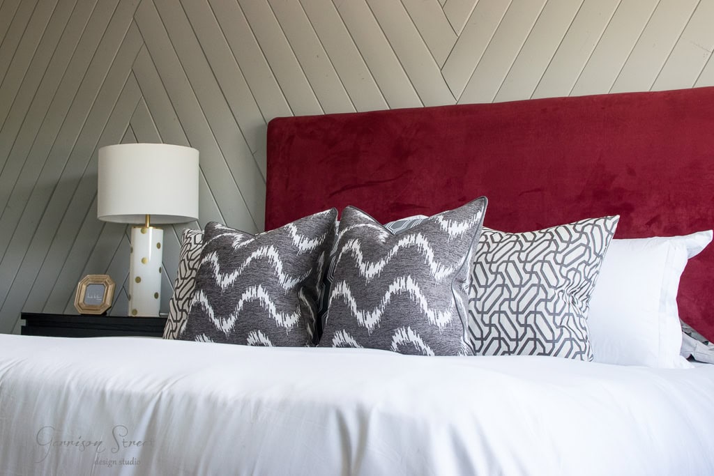 A modern bedroom features a bed with a red upholstered headboard and white bedding. Two patterned gray pillows, one with chevrons and one with wavy lines, are in front of the headboard. A white lamp sits on a black nightstand.