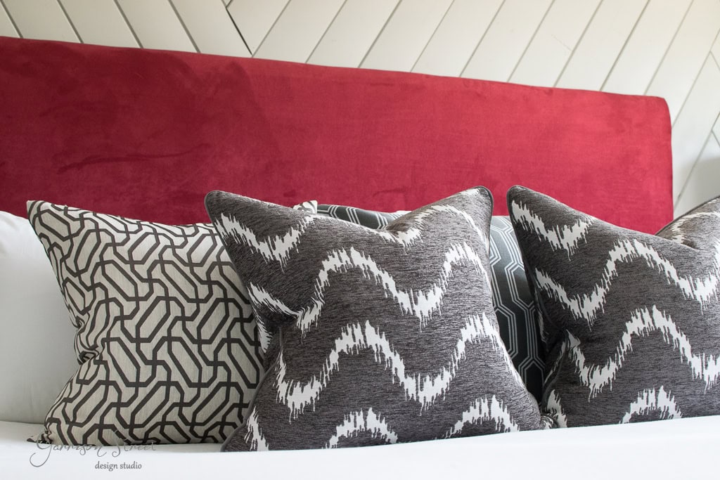 A bed with a red upholstered headboard features decorative pillows in various patterns, including zigzag and geometric designs, in shades of black, white, and gray.