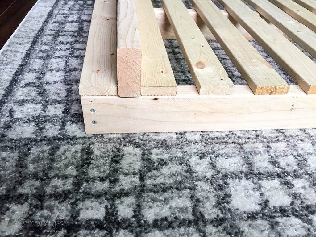 Close-up of a wooden bed frame with light-colored slats, resting on a gray and white patterned rug. The focus is on the corner of the frame, showing the clean lines and simple design.