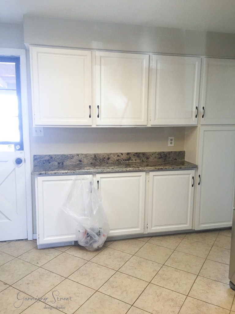 A kitchen with white cabinets and a granite countertop. A garbage bag hangs on a handle. The floor has beige tiles, and there's a door on the left.