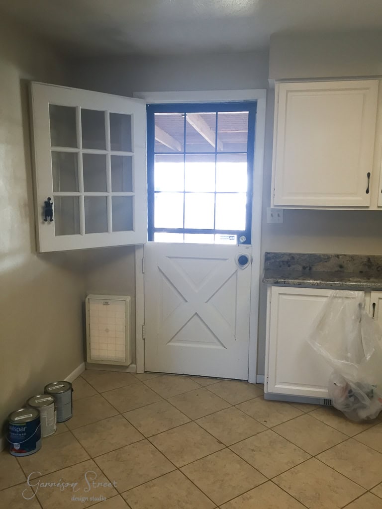 A kitchen with a half Dutch door partially open, and a window in the top half of the door. The room has beige tiled flooring, white cabinets, a granite countertop, and a few paint cans on the floor.