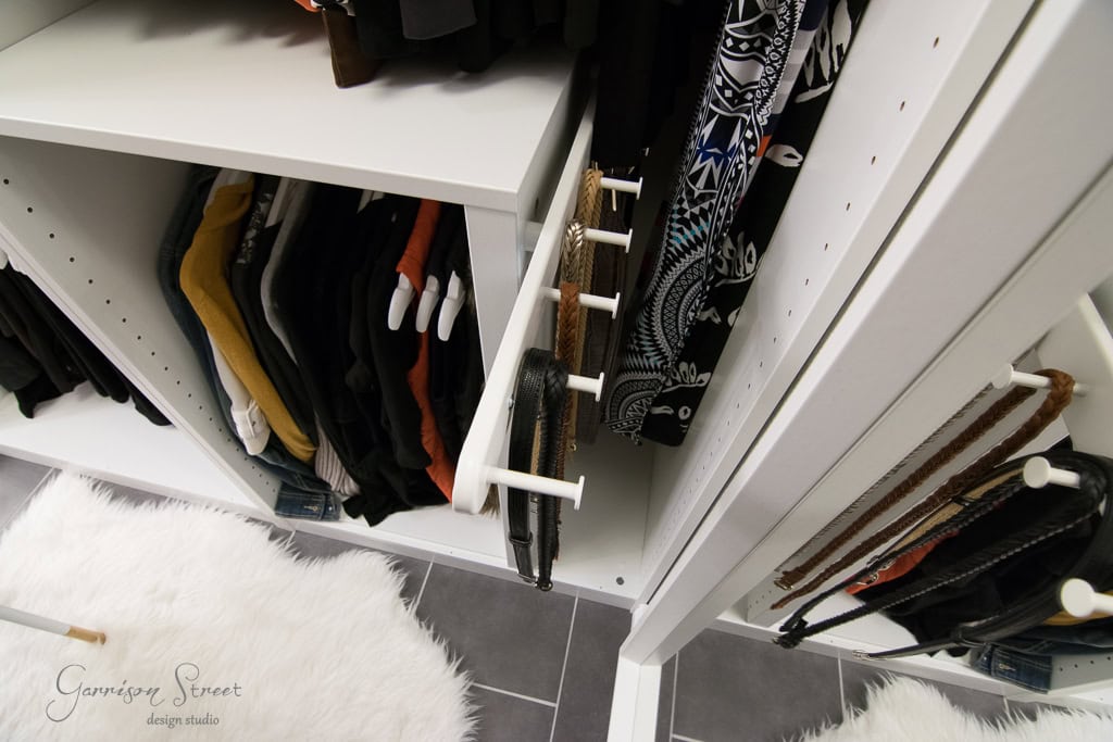 A neatly organized IKEA closet featuring hanging clothes, a pull-out rack with belts, and a white fuzzy rug on the floor. This walk-in design is clean and modern, with white shelves contrasting against dark floor tiles.
