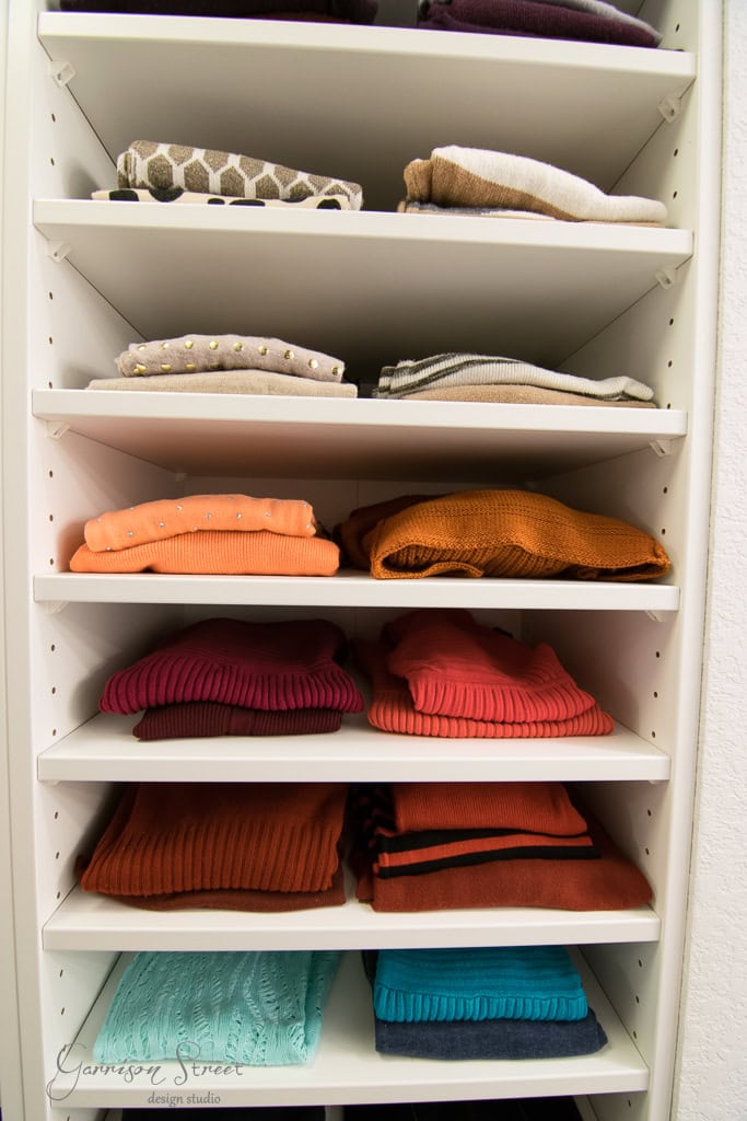 A spacious walk-in closet with neatly folded sweaters in an array of colors like grey, beige, orange, pink, and turquoise, all perfectly organized on sleek white IKEA shelves.