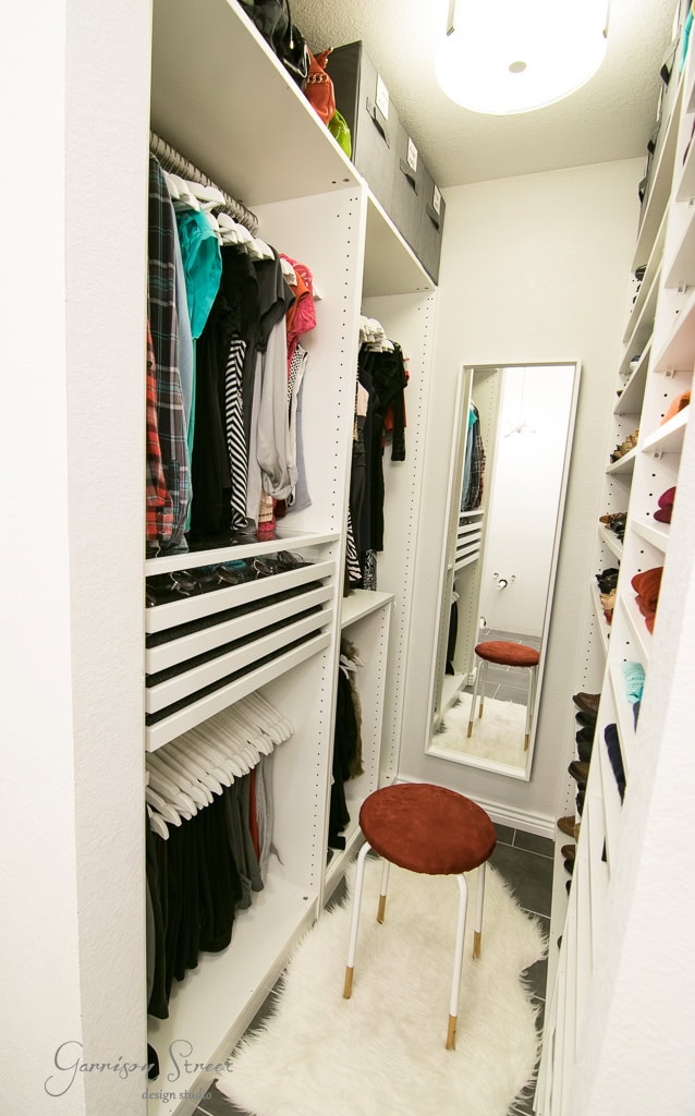 A narrow IKEA walk-in closet features white shelves and hanging clothes, with a full-length mirror at the end. A small round stool with a brown seat and a white fluffy rug grace the center. The space is brightly lit with ceiling lights.