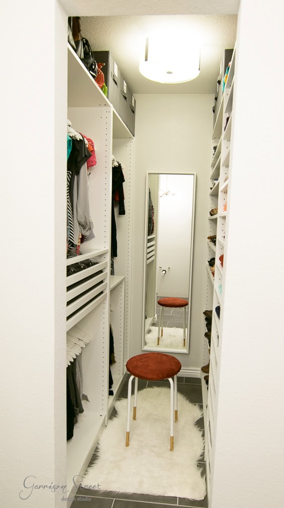 This IKEA-inspired walk-in closet features sleek white shelves and clothes neatly hanging on both sides. A full-length mirror graces the far end, while a small red cushioned stool rests on a plush white fur rug beneath a stylish ceiling light.