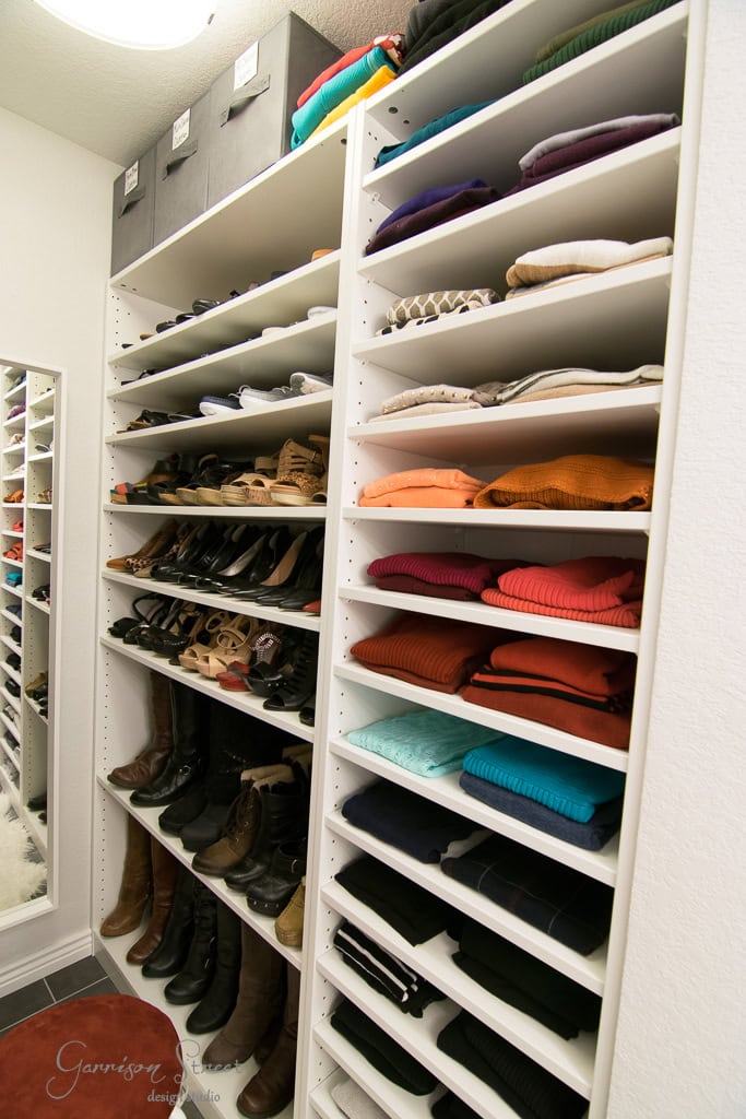 A beautifully arranged walk-in closet boasts white IKEA shelves, storing an array of colored sweaters, neatly folded clothes, and a curated collection of shoes and boots. Labeled boxes adorn the top shelf, while a mirror reflects the tidy space.
