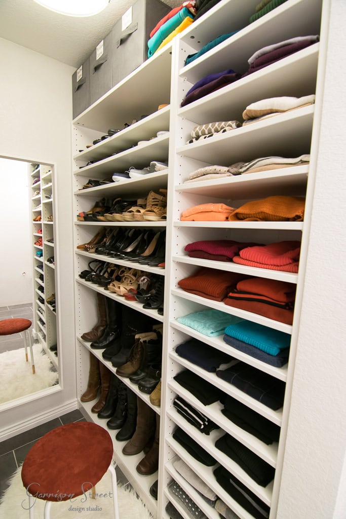A well-organized IKEA closet features shelves filled with neatly folded clothes and rows of shoes. A large mirror stands to the left, while a small red stool is placed in front. Overhead storage boxes are lined up on top, completing this efficient walk-in space.