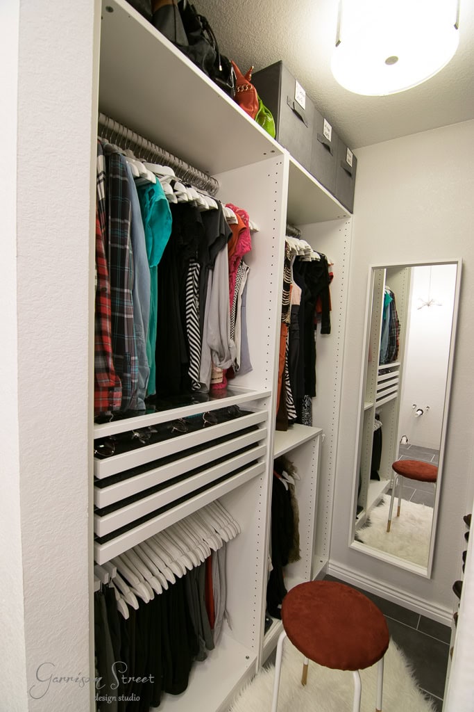The walk-in closet, reminiscent of an IKEA display, boasts white shelves with hanging clothes, folded items, and storage boxes. A full-length mirror graces the wall beside a small stool with a brown cushion resting on a plush white rug.