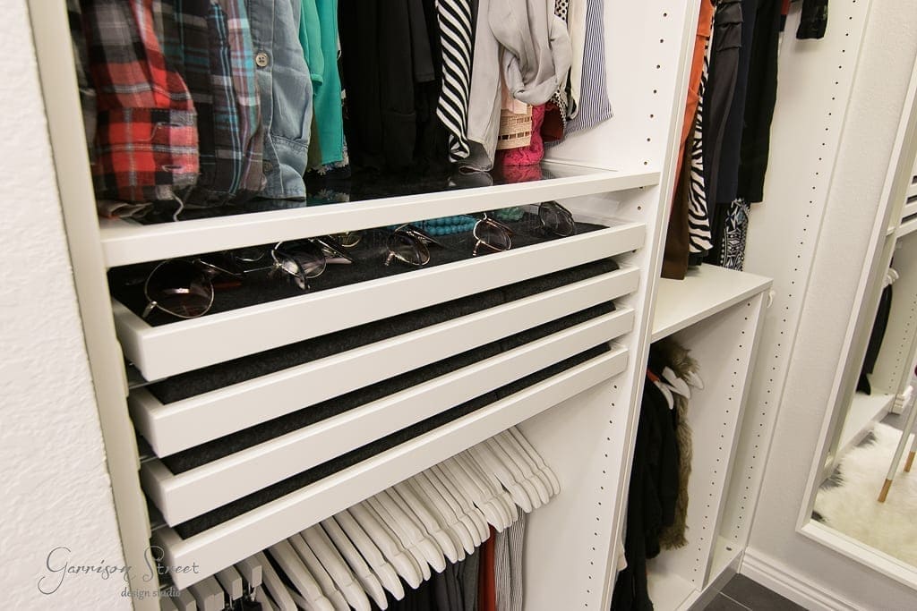 Home A neatly organized IKEA walk-in closet features folded shirts on shelves, sunglasses displayed in pull-out drawers, and hangers holding various clothes. A mirror is visible on the right.