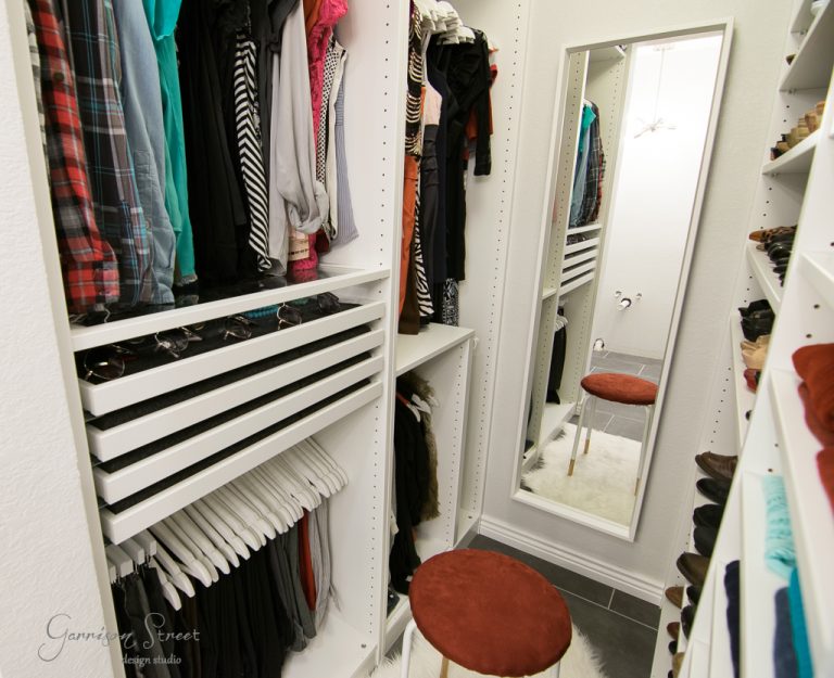 A small, organized walk-in closet with shelves and hanging clothes on both sides. An IKEA full-length mirror leans against the back wall, and a round stool with a red cushion is positioned on a white rug in the center.