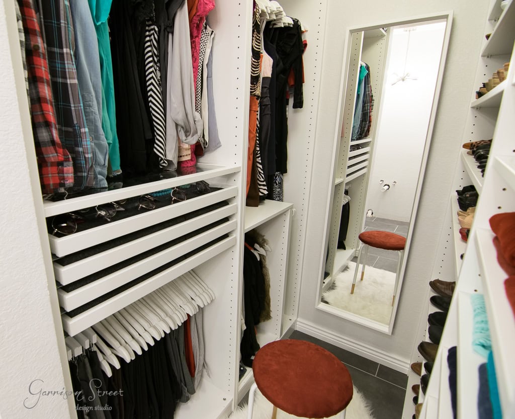 A small, organized walk-in closet with shelves and hanging clothes on both sides. An IKEA full-length mirror leans against the back wall, and a round stool with a red cushion is positioned on a white rug in the center.
