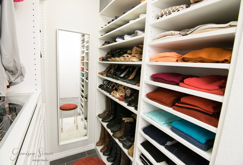 A well-organized IKEA walk-in closet features shelves brimming with folded sweaters and multiple rows of shoes, from boots to heels. A tall mirror leans against the white wall, perfectly paired with a charming red stool.
