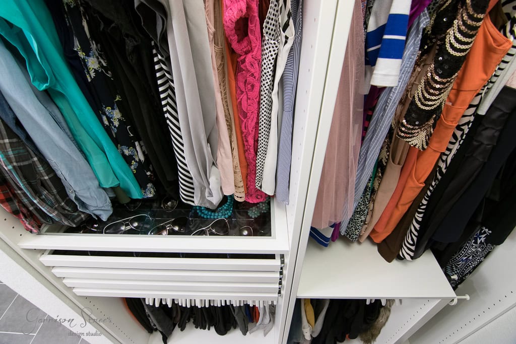 A walk-in closet features neatly arranged colorful clothing, such as shirts, dresses, and blouses on hangers. An IKEA-inspired drawer below houses sunglasses, while another section with more garments is visible on the right.