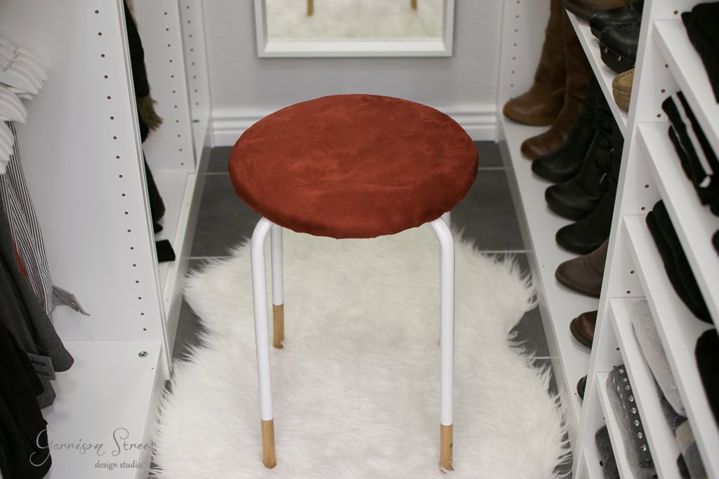 A small, round IKEA stool with a red cushion and white legs sits on a fluffy white rug in the walk-in closet. The space boasts organized shelves filled with clothes and shoes, complemented by a mirror on the back wall.
