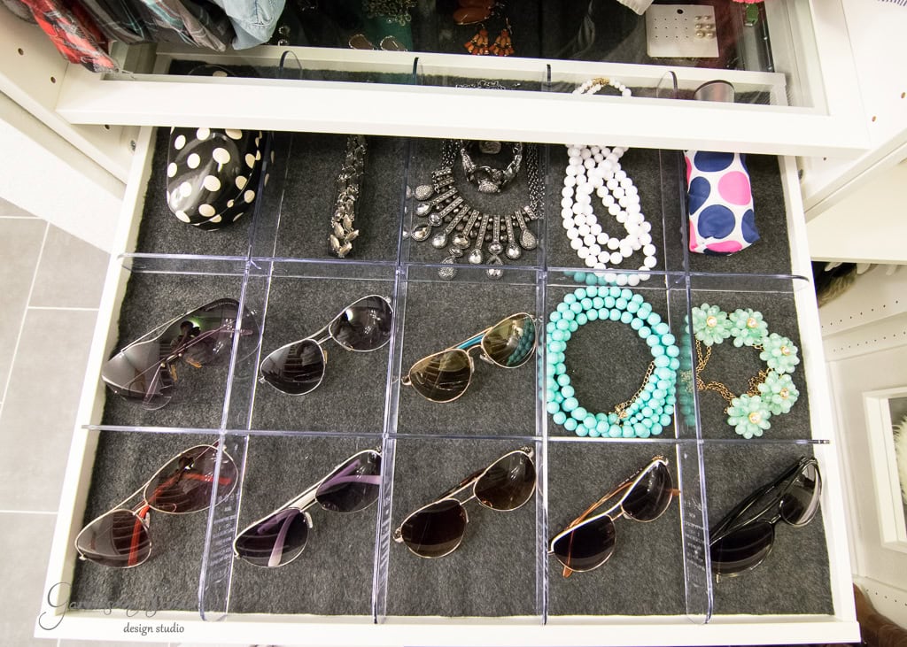A walk-in closet reveals a drawer with organized compartments displaying sunglasses, necklaces, bracelets, and an IKEA-patterned glasses case. The drawer is lined with dark fabric, neatly arranging the accessories in individual sections.