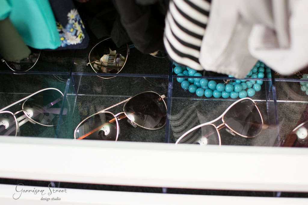A walk-in closet houses a meticulously organized drawer with several pairs of sunglasses featuring reflective lenses. Above them, clothes and a turquoise beaded necklace add a touch of charm, creating an atmosphere reminiscent of an IKEA showroom.