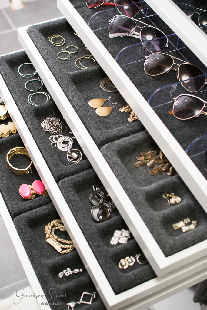 An IKEA jewelry organizer features multiple trays for accessories like rings, bracelets, sunglasses, and earrings. Perfectly suited for a walk-in closet, it keeps items neatly arranged in dark fabric-lined compartments.